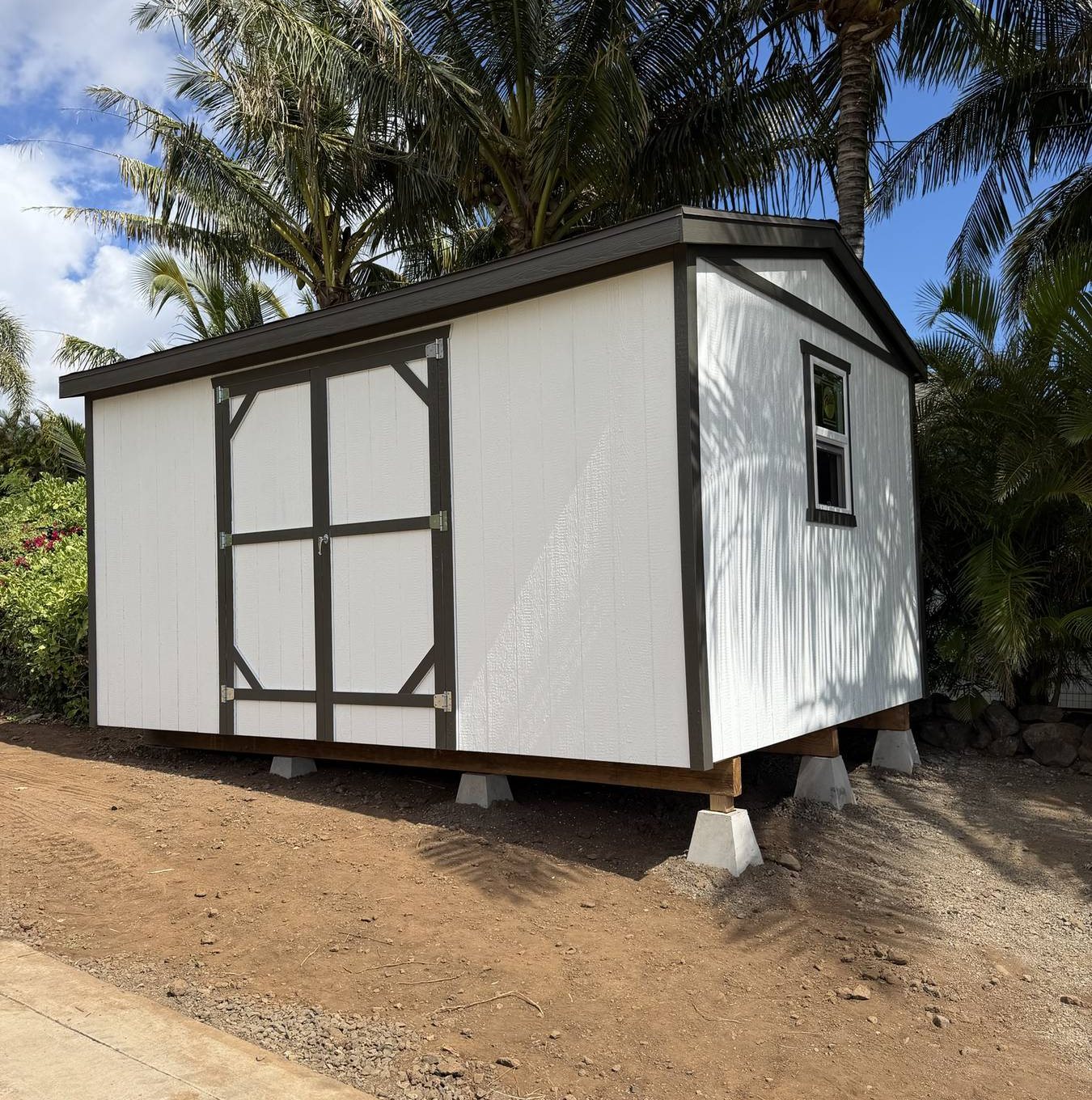 White custom storage shed with black trim and a barn door, set on piers under palm trees on Maui